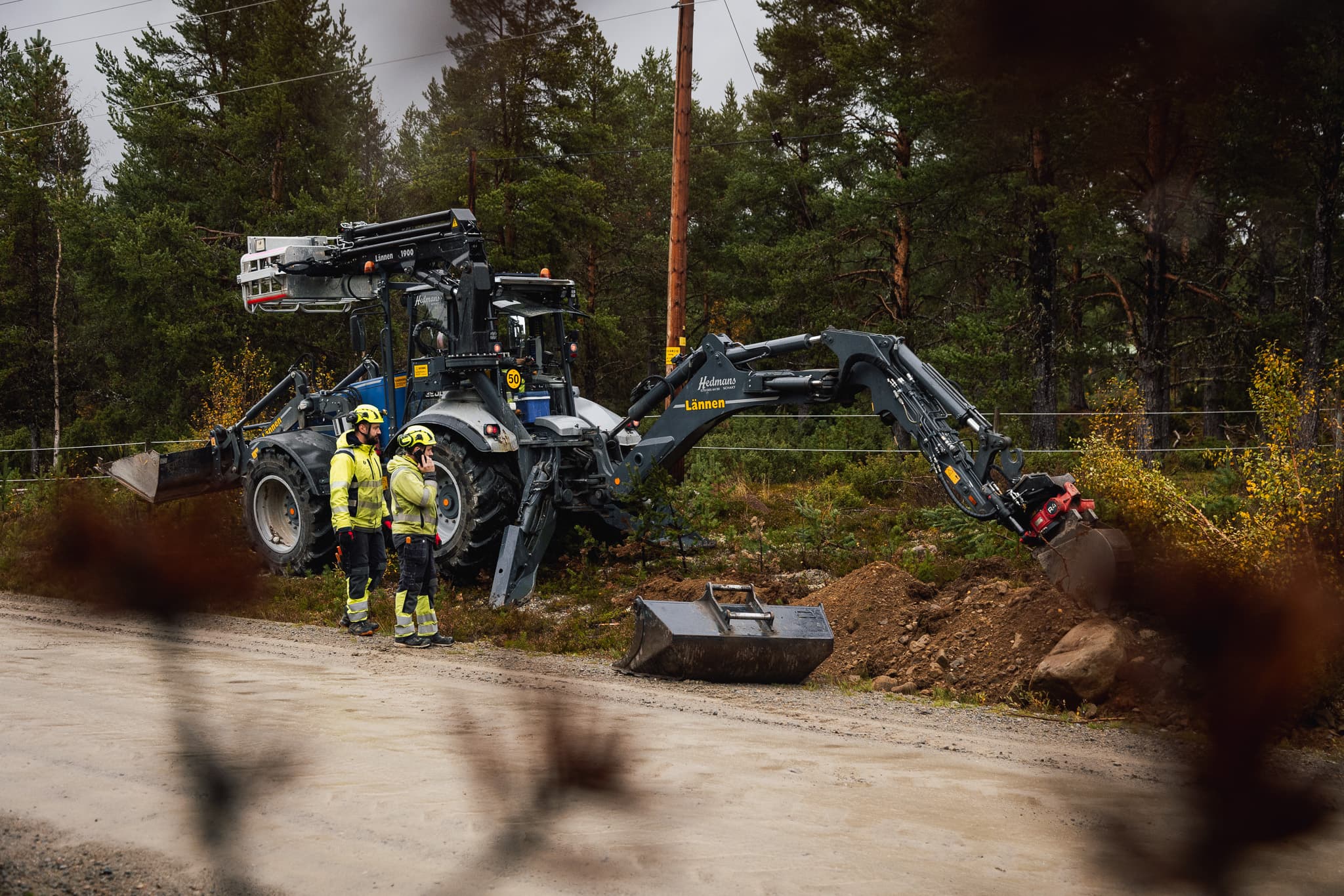 Traktorgrävare med manskapskorg som arbetar vid elledningar under utbyggnad av elnät och fjärrvärme i södra Jämtland av BTEA.
