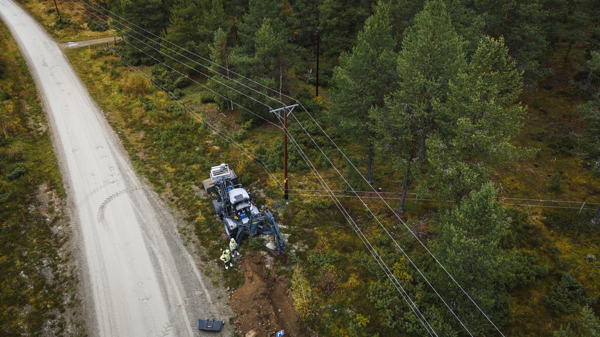 Grävarbete invid kraftledningar längs en väg fotat ovanifrån